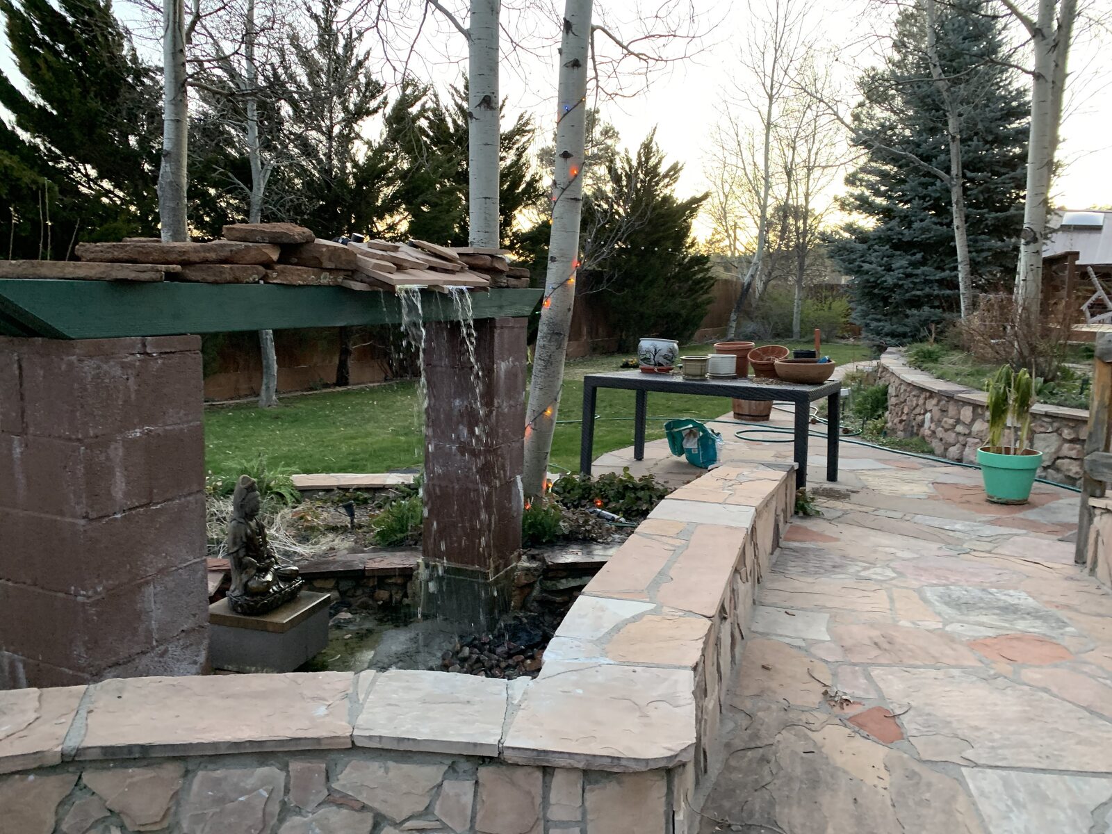 A hand-built garden waterfall falling into a koi pond at dusk, with a stone Kwan-Yin figure beside it